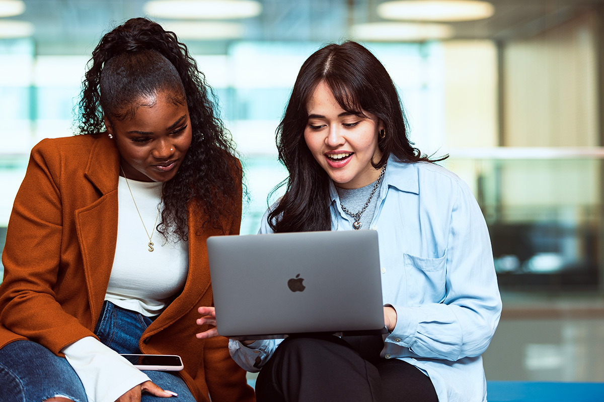 two students sitting in a campus lobby looking at a laptop screen