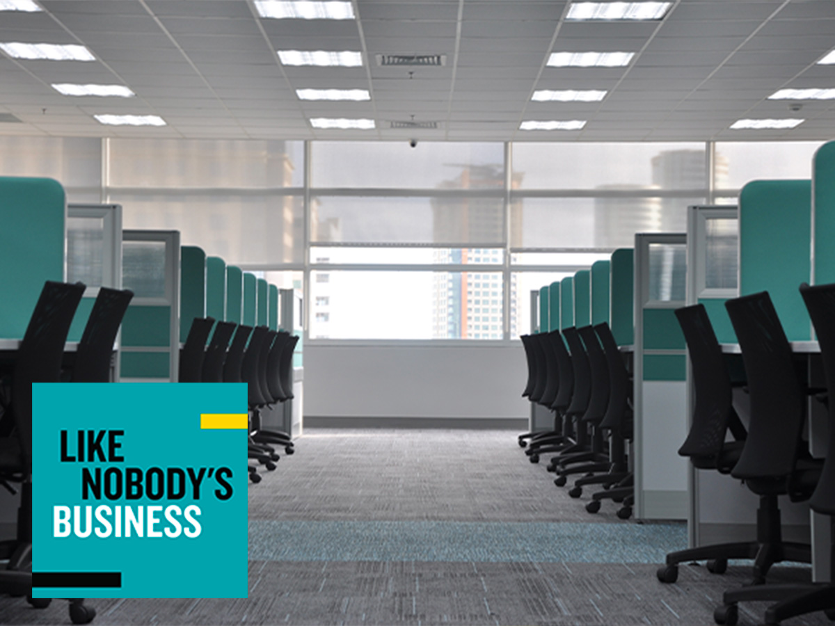 An empty office with black chairs, blue dividers at each desk and grey carpet. There is a window at the end of the hallway and the Like Nobody's Business in the bottom left corner, with a teal background and black and white block text. 