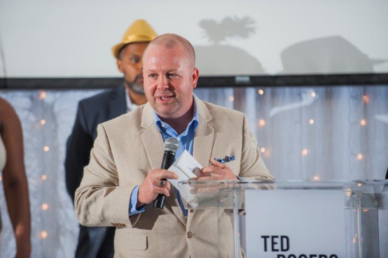A middle aged man in a beige suit reading off a slip of paper the winner of an award at a ceremony