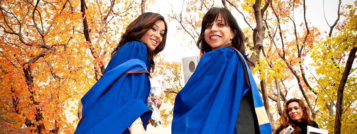 Two female students in their graduation gowns looking behind them for a picture under bright orange autumn leaves