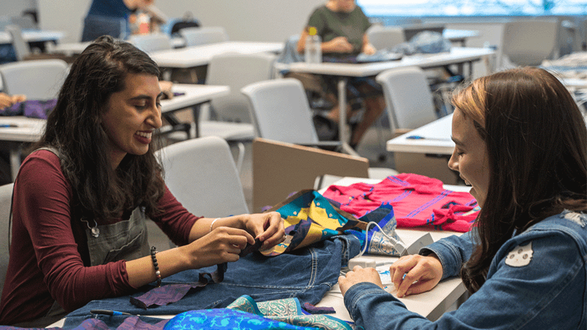 Two students sit at a table sewing fabric