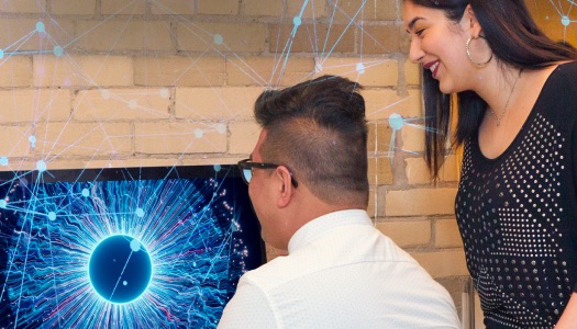 Two students collaborate on a project featuring a glowing, neural-network-like graphic on a computer screen.