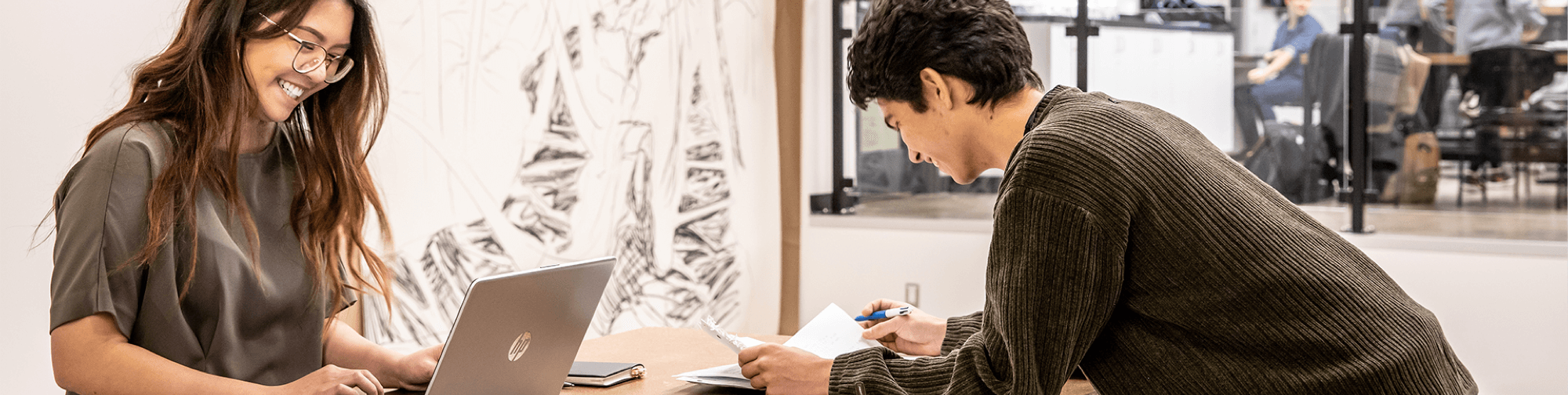 Student smiling with laptop in front of a student holding a notebook and a pen