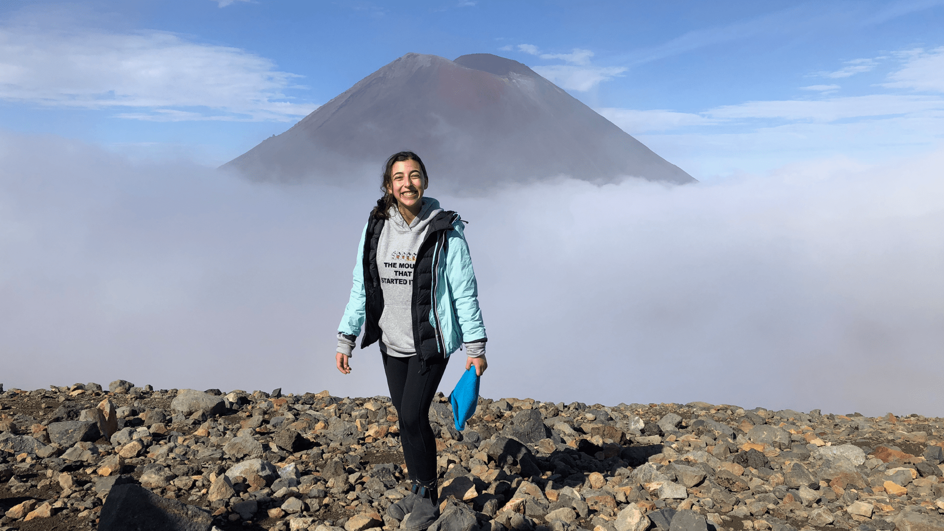 smiling girl in a light blue jacket and grey hoodie on top of a mountain with low clouds and scenic mountaintop view
