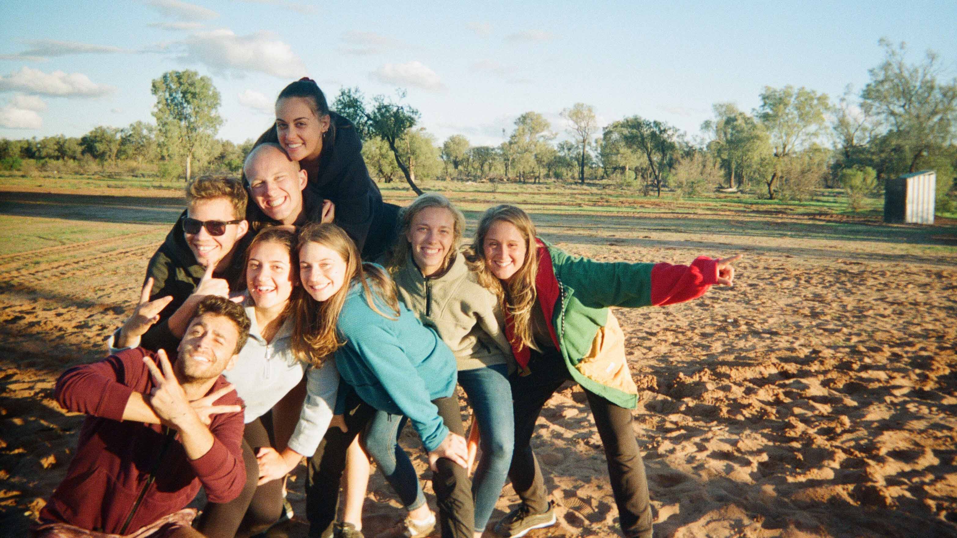 Group of students hugging on a sand beach with trees in the back