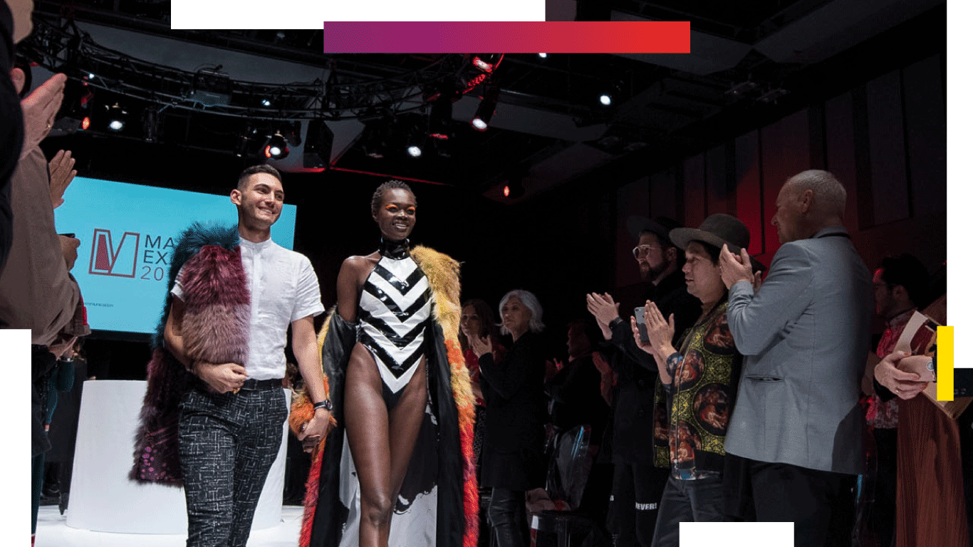 A model and designer walk the runway at the year end fashion show. Model is wearing a body suit with a black and white chevron pattern that points downwards, as well as a cape they are wearing off their left shoulder