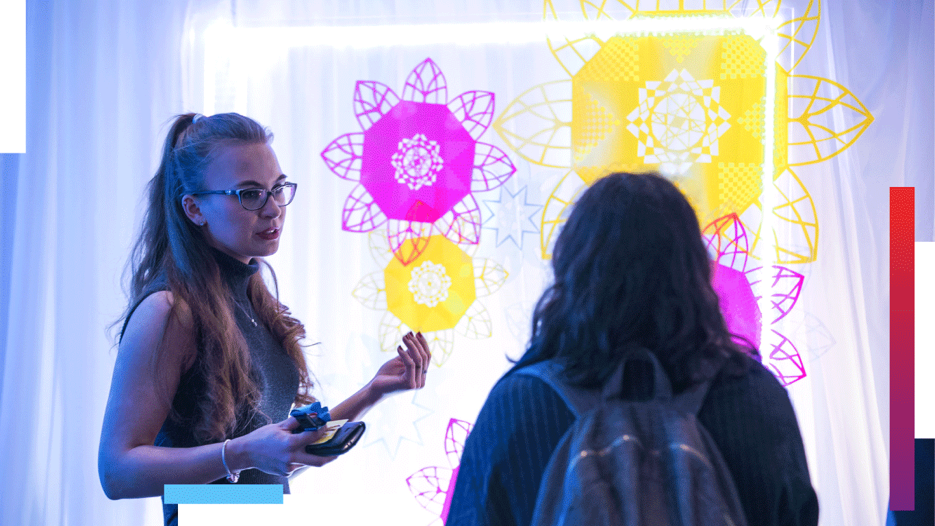 Two students stand in front of a bright lit up art display of abstract flower shapes