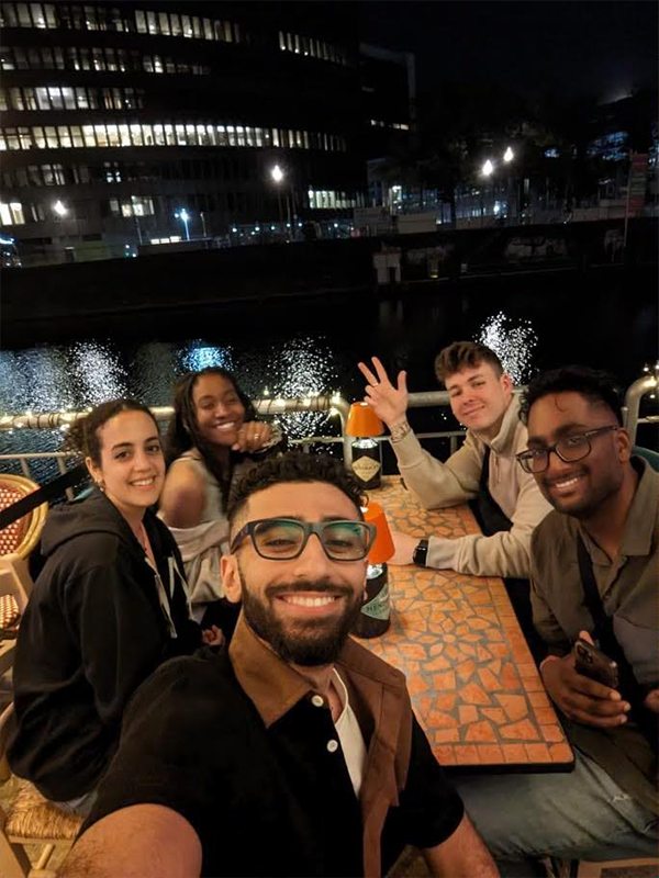 A group of students sitting around a table outside pose in front of a river at night.