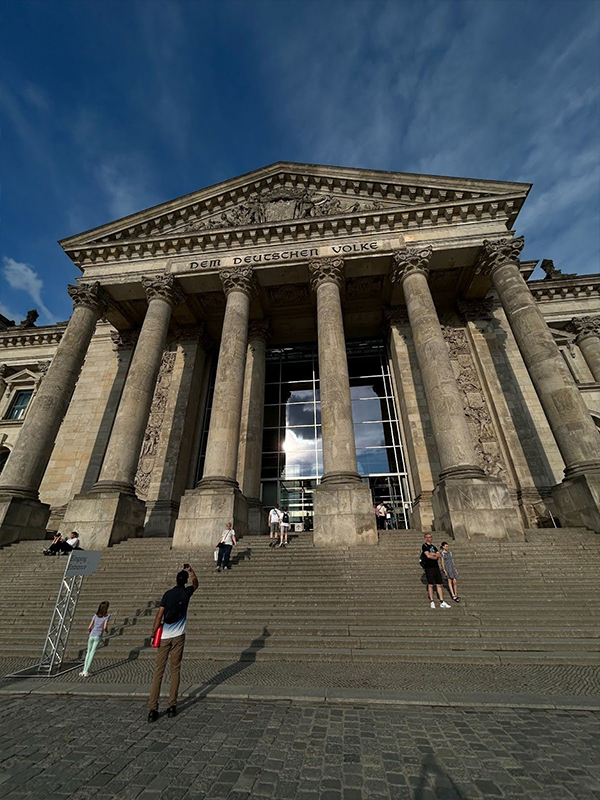 A group of people stand sporadically on the stairs of a historic building with six large pillars marking its entrance.