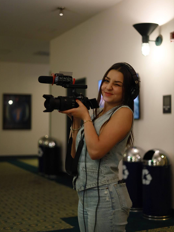 A woman holds a camera and winks at someone taking her picture.