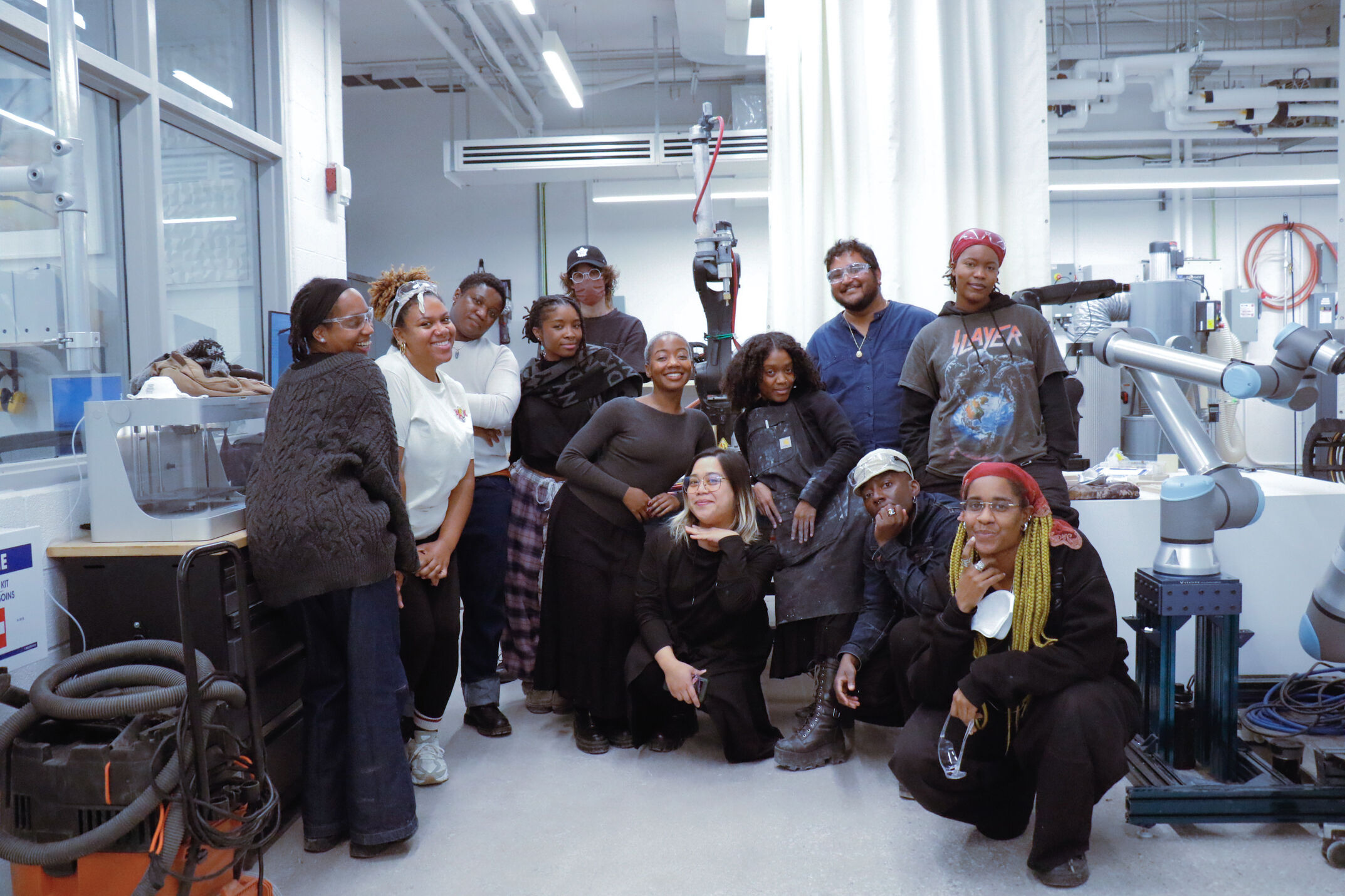 A group of 12 people pose and smile together inside a brightly lit fabrication lab. They are standing and crouching around a light blue and silver industrial robotic arm. In the background, there is various lab equipment, including a 3D printer enclosure, a shop vac, and exposed ceiling pipes.