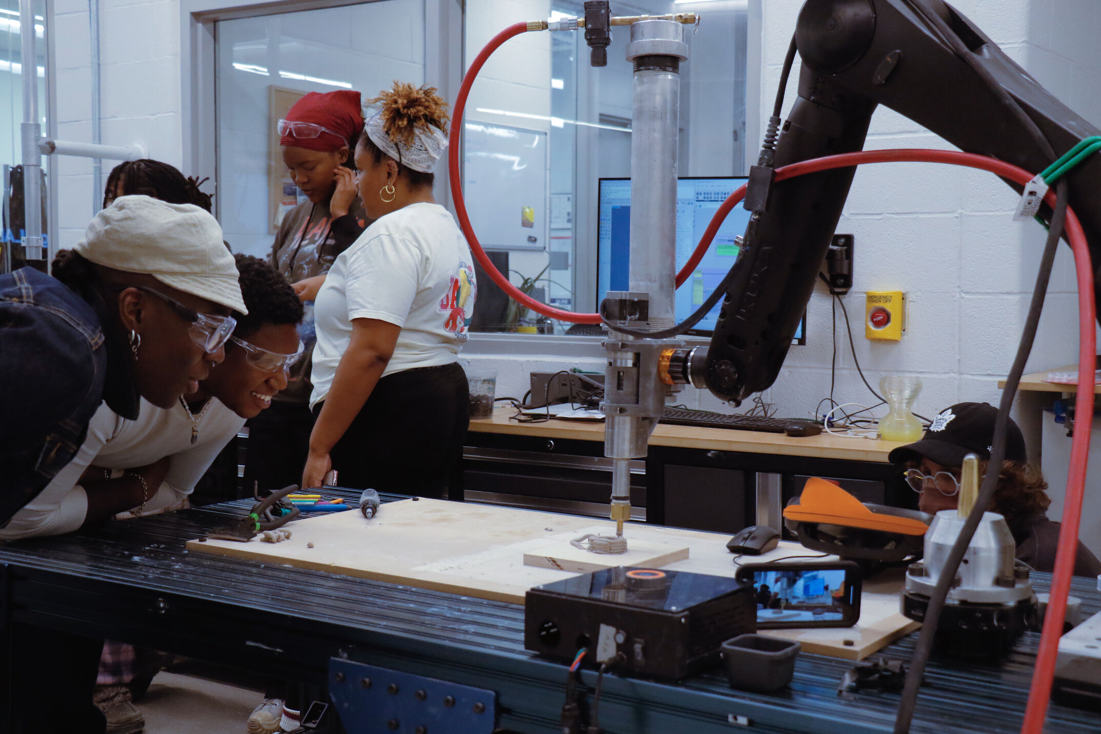 Three people, two wearing safety glasses, lean over a black metal workbench, watching intently as a robotic arm with a silver nozzle prints a small clay object. In the background, three other participants are chatting. The lab is brightly lit with a window and a computer monitor visible.