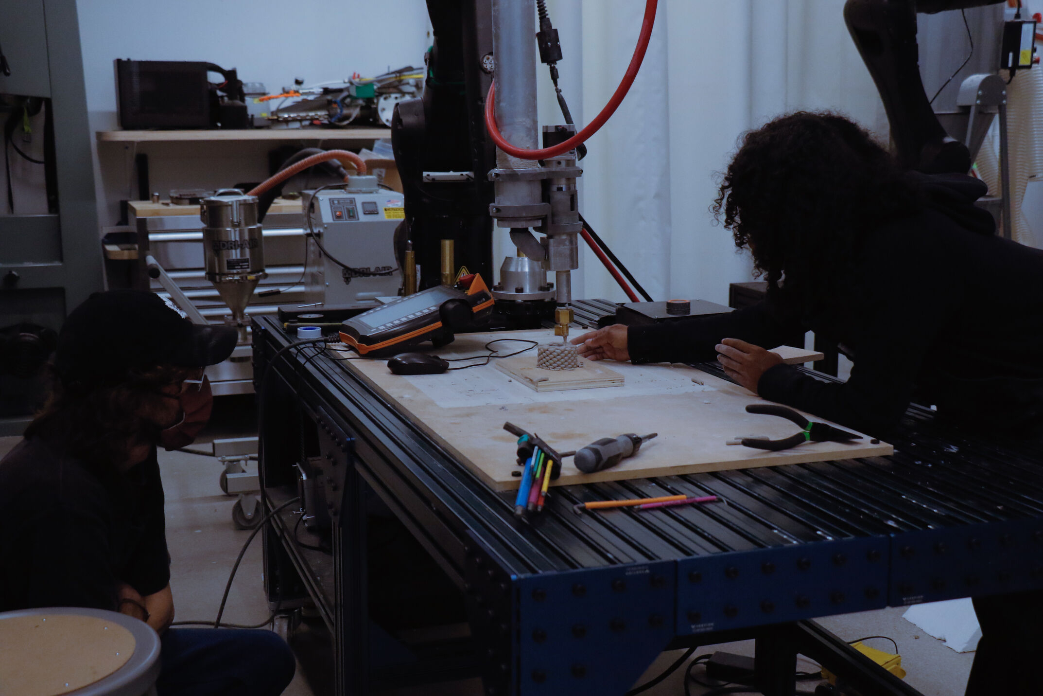 In a dimly lit lab, a person with long curly hair leans over a workbench, using their hands to inspect or adjust a small, textured clay object that has just been printed by the large robotic arm above it. Various tools, including pencils and pliers, are scattered on the workbench surface. Another person wearing a black cap sits beside the table, watching the process. 
