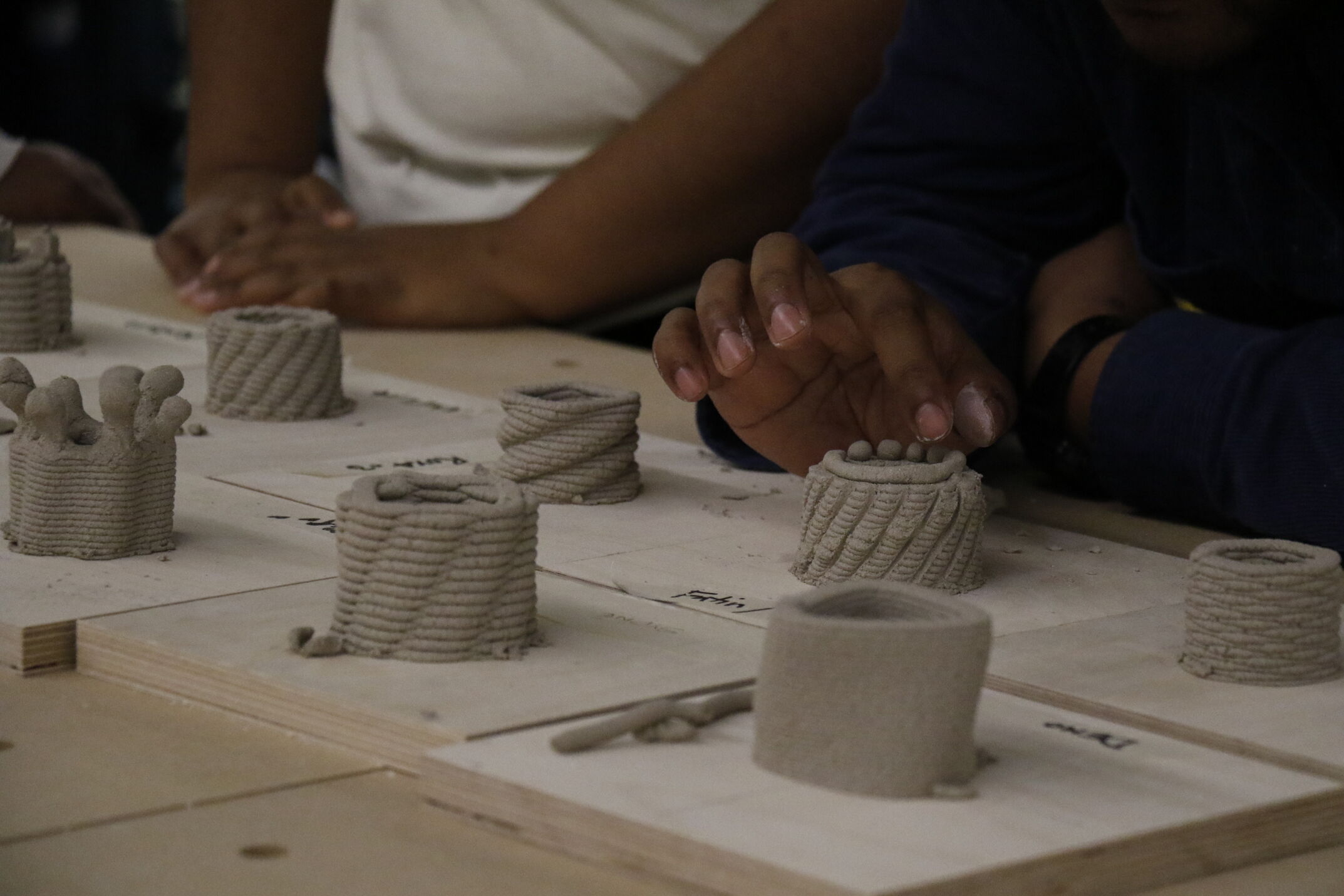 A close-up shot of several small, 3D-printed clay vessels sitting on square wooden boards. The vessels feature a distinctive, layered coil texture. A person's hand reaches into the right side of the frame to gently touch the scalloped top edge of one of the wet clay pieces. Another person's arms are visibly resting on the table in the blurred background.