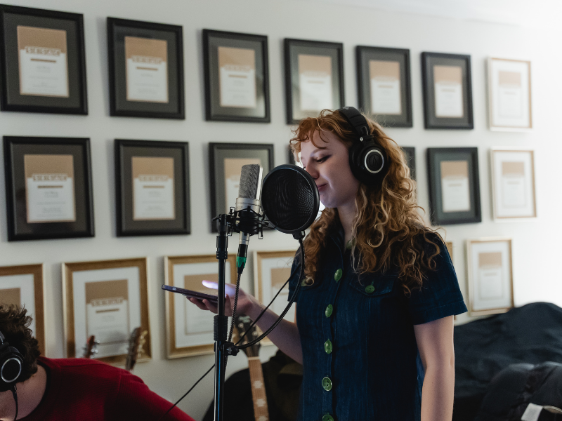 A student is wearing headphones, holding her phone up and singing into a mic in what looks like a recording studio.