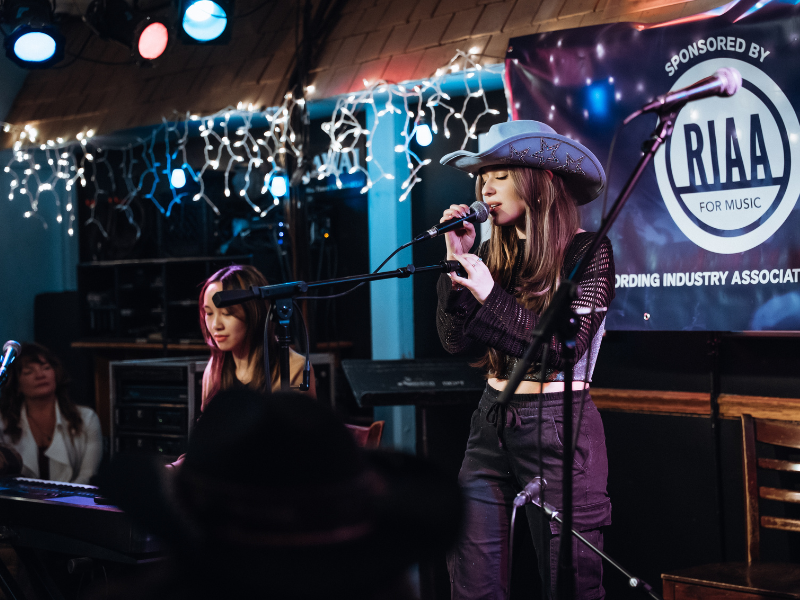 Student is wearing a cowboy hat, singing into a mic on a stage. 