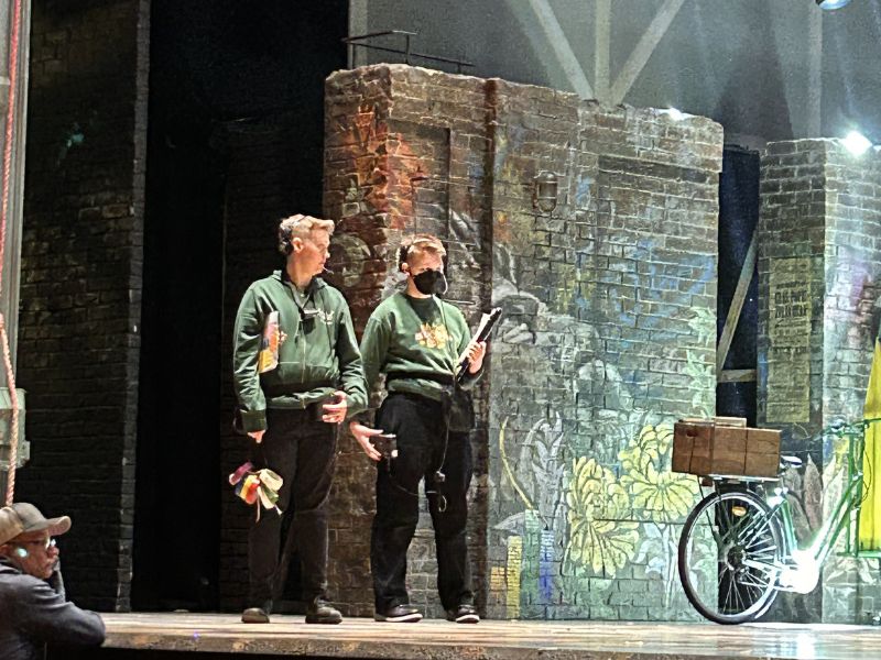 In matching green hoodies, alumni Anna and Ciaran stand on stage during a reherasal for & Juliet. Each person is holding backstage equipment such as tape and a clipboard.