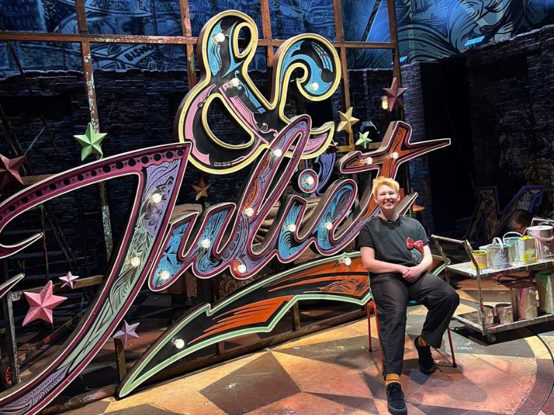 Ciaran Brenneman is seated in front of the & Juliet sign smiling for a photo backstage before the hanging of the sign on stage.