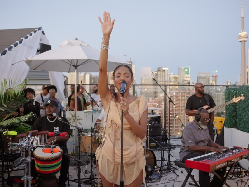 Naomi is standing in front of a microphone, performing at a Toronto rooftop music event. The CN Tower is in the background and she is surronded by a band. 