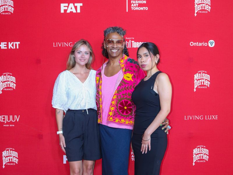 Three people pose together on a red carpet in front of a bright red step-and-repeat backdrop featuring Fashion Art Toronto and sponsor logos. The person in the center wears a vibrant pink floral jacket over a pink top and a long dark skirt, smiling at the camera. On the left, a person in a white blouse, dark shorts, and blue sandals stands with a relaxed pose. On the right, a person in a fitted black sleeveless dress and black sandals poses with one hand in a pocket. 
