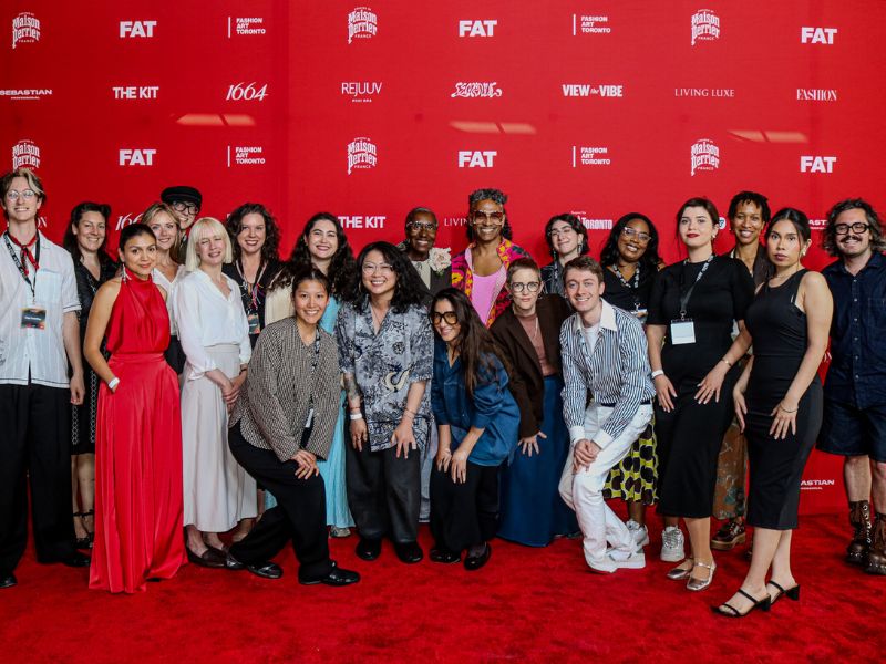 Group photo of approximately 18 people posing on a red carpet in front of a bright red step-and-repeat backdrop featuring Fashion Art Toronto (FAT) and sponsor logos. The group is dressed in a mix of formal and creative attire, smiling and facing the camera.