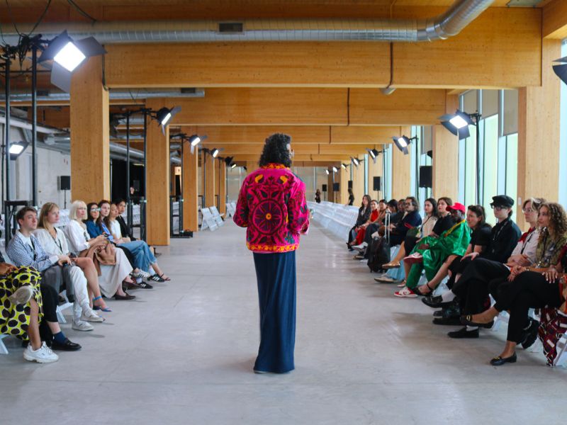 A person wearing a bright pink embroidered jacket and a long dark skirt stands at the center of a fashion runway, facing away from the camera and addressing attendees seated on both sides. The spacious venue features exposed wood beams, large windows, and professional runway lighting. Guests in a variety of colorful and contemporary outfits watch from white folding chairs.