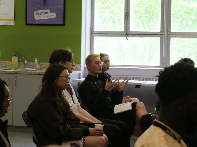 Several people sit in a classroom discussion, listening as one participant speaks and gestures with their hands while holding a notebook on their lap. Other attendees sit nearby, some taking notes or holding cups. Large windows let in daylight, and a sign reading “Fashion at the Creative School” hangs on the green wall above a counter and sink in the background.