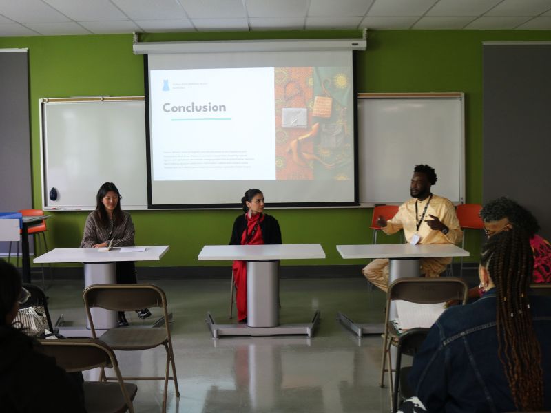 A panel discussion takes place in a classroom. Three speakers sit at separate tables at the front of the room while audience members listen from folding chairs. Behind them, a projector screen displays a presentation slide titled “Conclusion,” and whiteboards line the green walls. One speaker gestures while talking, and the others look on attentively.