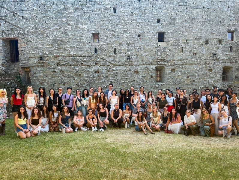 Wide-shot group photo of about 60 students in front of a historic fortress in Italy.