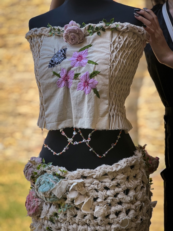 A mannequin dressed in a cream two-piece garment with smocking, floral embroidery, crochet details, and fabric flowers.