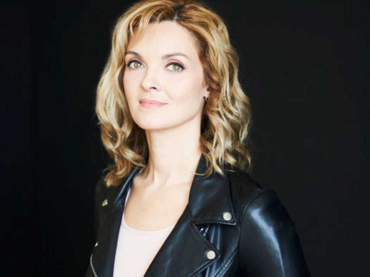 A headshot of Ramona Pringle against a black background. She looks at the camera with a slight smile and has blonde hair with light curls. Ramona is wearing a black leather jacket.