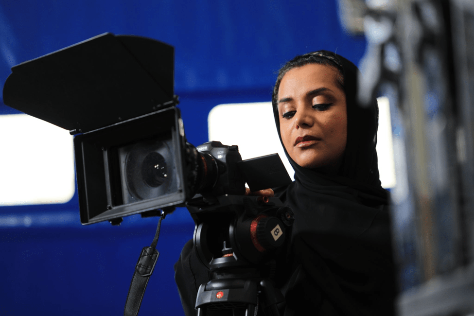 Al Khaja wears all black and looks at the screen of a black camera on a tripod.