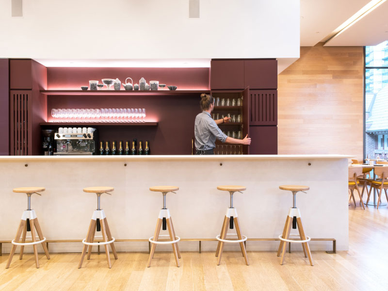 Picture of the bar at Clay restaurant. The back counters are painted a deep aubergine and the bar counter is white. In front of the counter are five potter stools. A server grabs a glass behind the bar