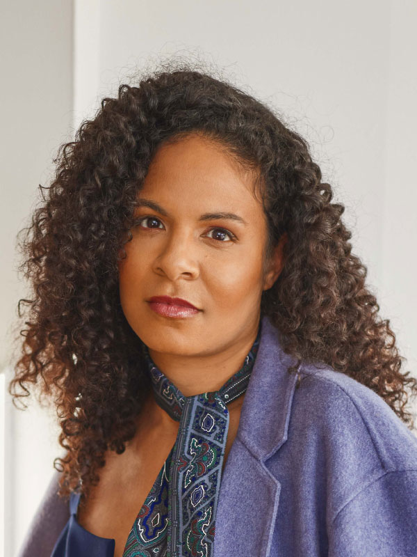 Portrait of Dyonne Fashina. Fashina wears a purple jacket and a blue patterned scarf. Her hair is dark and curly and she looks into the camera
