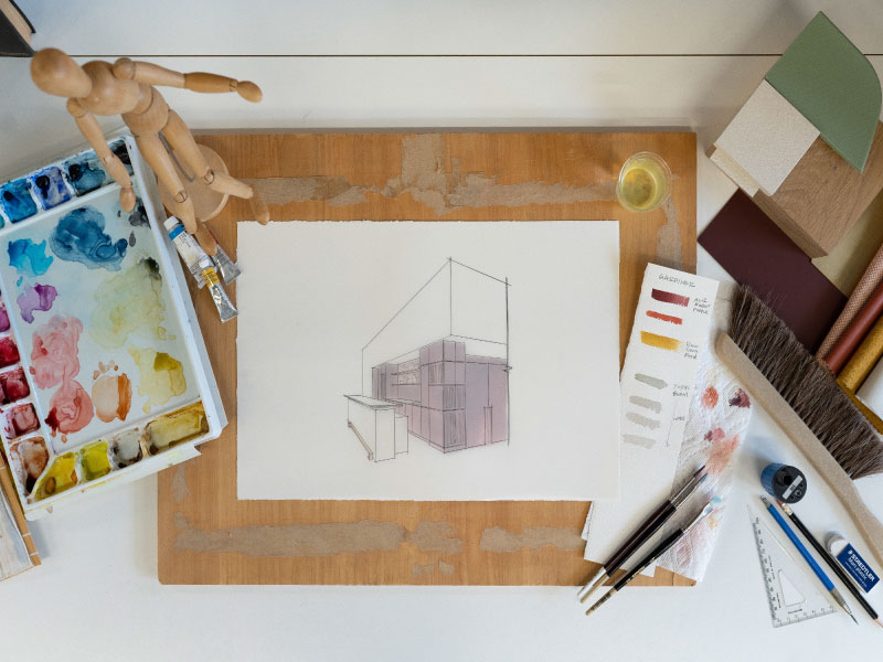 A mockup of the bar design is drawn on a piece of white paper and filled in with purple watercolor. To the left there is a paint palette and to the right, pencils and paint swatches