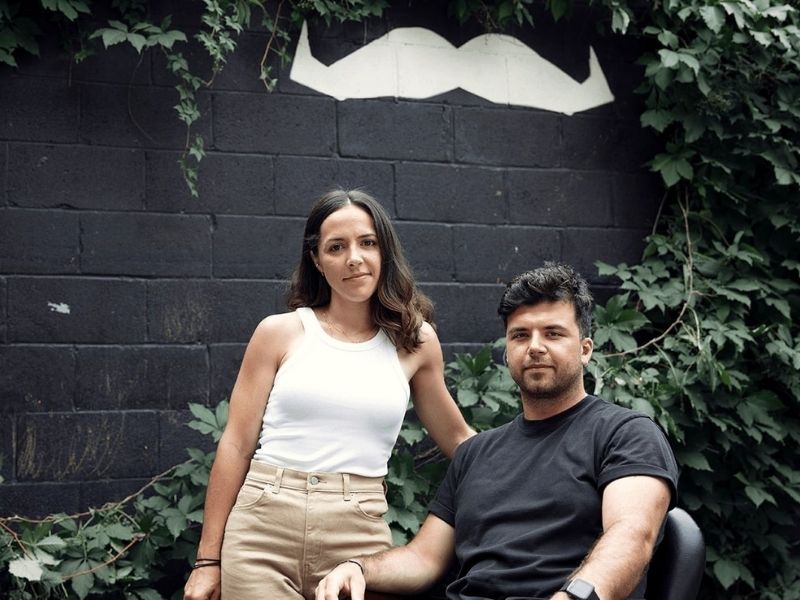 The co-founders of Hello Jack posing in front of a black wall with a white moustache painted on it and green foliage growing up the wall