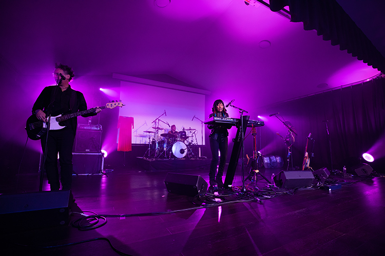 Woman sings into a mic while playing keyboard. Man plays electric guitar on stage with her