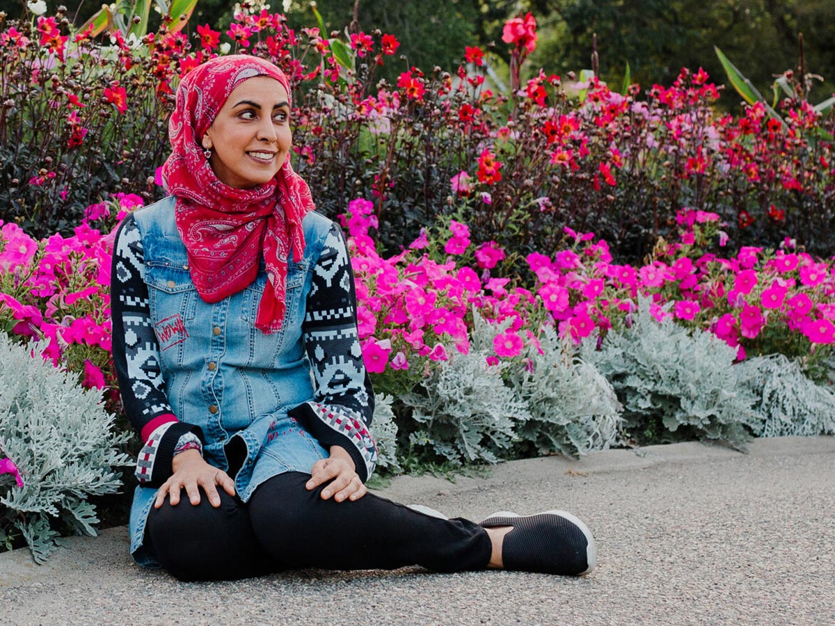 Zarqa Nawaz wearing a bright pink headscarf sitting in front of a flower bush