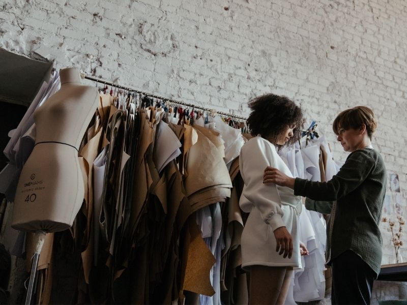 Woman gets fitted for a white dress by another woman in front of a rolling rack filled with hanging patterns and near a judy doll