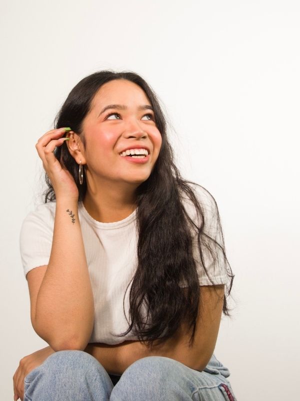 Young woman sits and looks up with one hand placed in her hair. She wears a t-shirt and jeans and is smiling