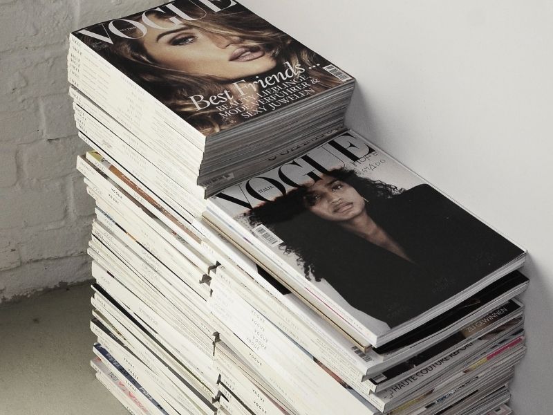 Two stacks of fashion magazines lay on a concrete floor and next to a white wall. Two Vogue magazine covers with women are visible