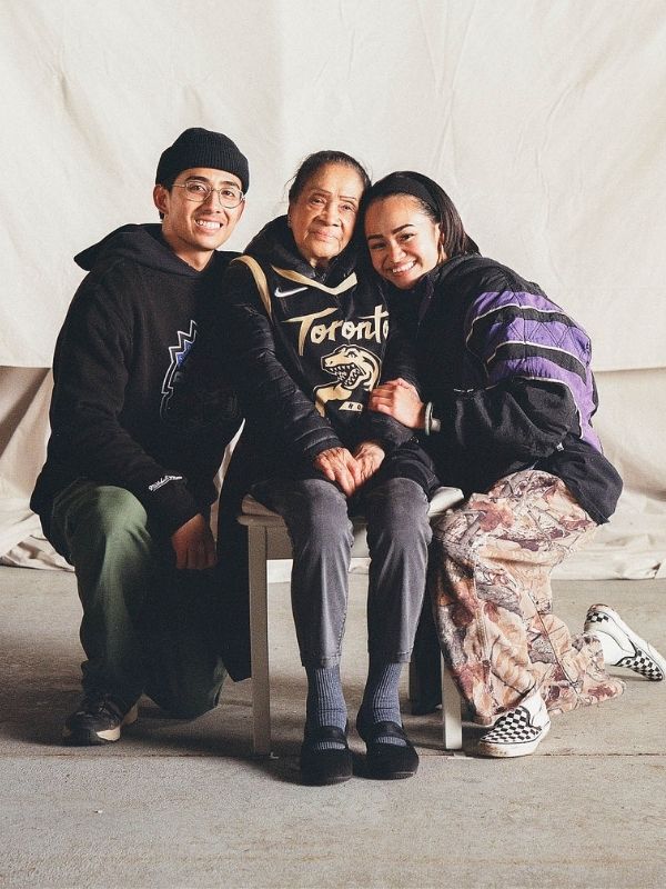 Young man kneels next to grandmother who sits on a chair and next to a young woman. All are wearing NBA Toronto Raptors branded apparel. 