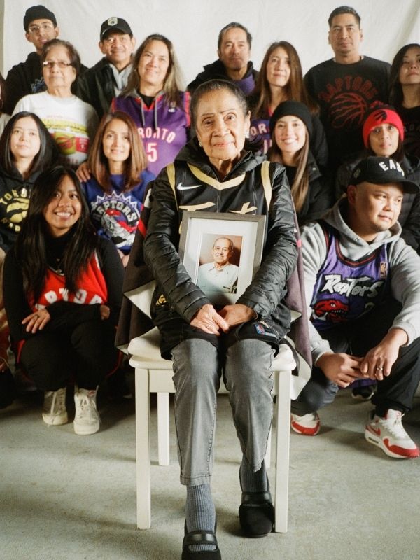 Senior woman sits in chair wearing a Toronto Raptors jersey, holding framed image of her husband. Behind her are posed many members of her family, all wearing braned apparel