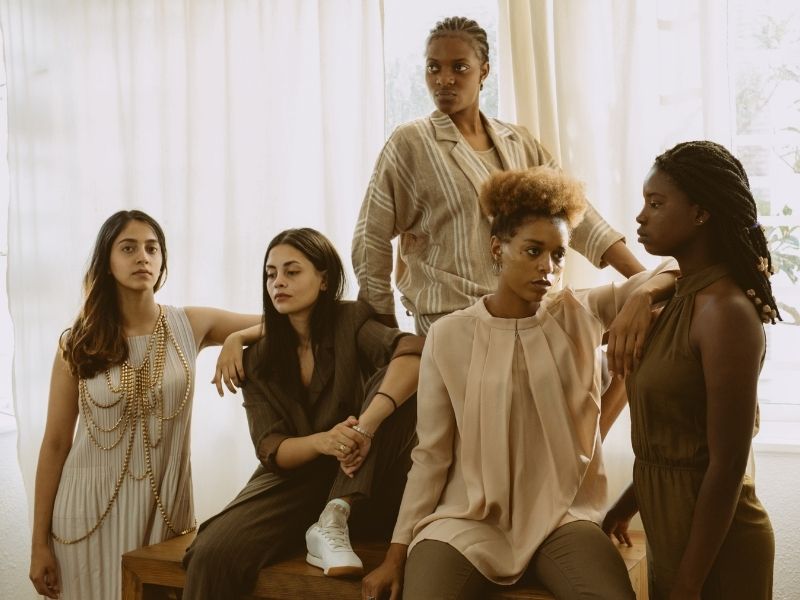 Five women models standing and sitting in various poses close together, wearing neutral tones