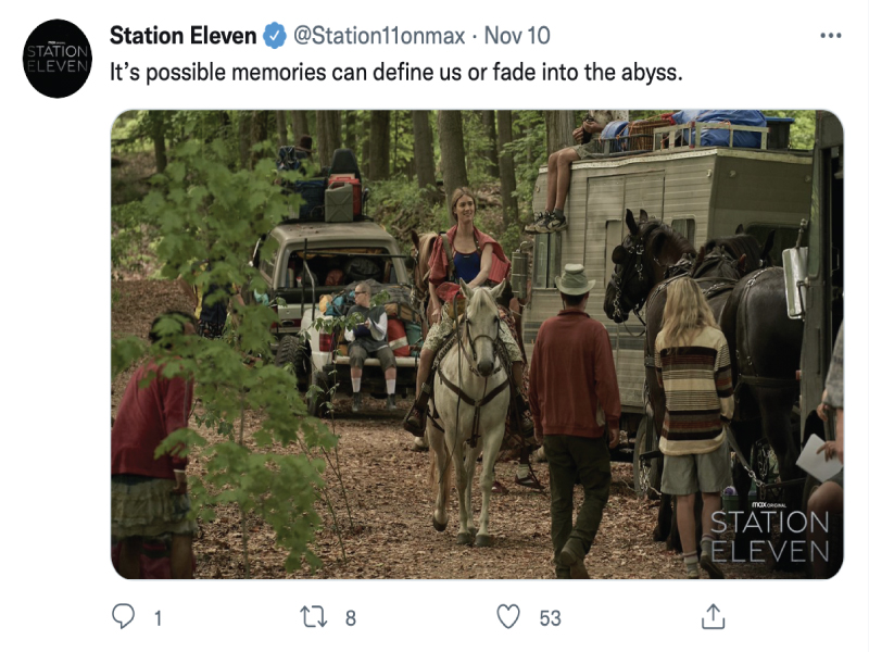 Image shows a group of people in the forest with trailers and vans packed with personal belongings. One woman rides a white horse towards the camera while everyone else looks away from the camera. 