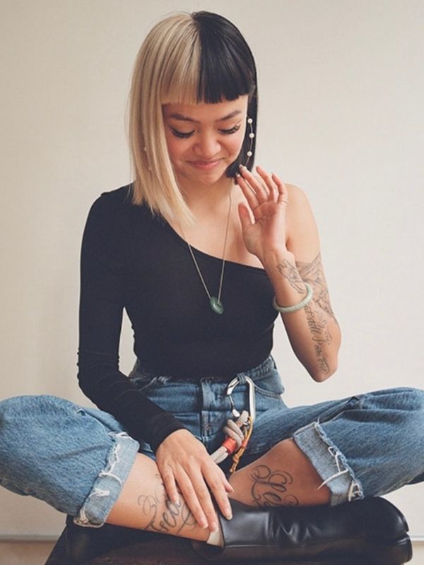 Young woman sitting cross legged and looking downwards with one hand near her hair which is dyed half blond, half black