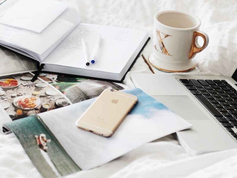 Laptop open on a bed next to a mug, notebook, pens, magazine an cell phone