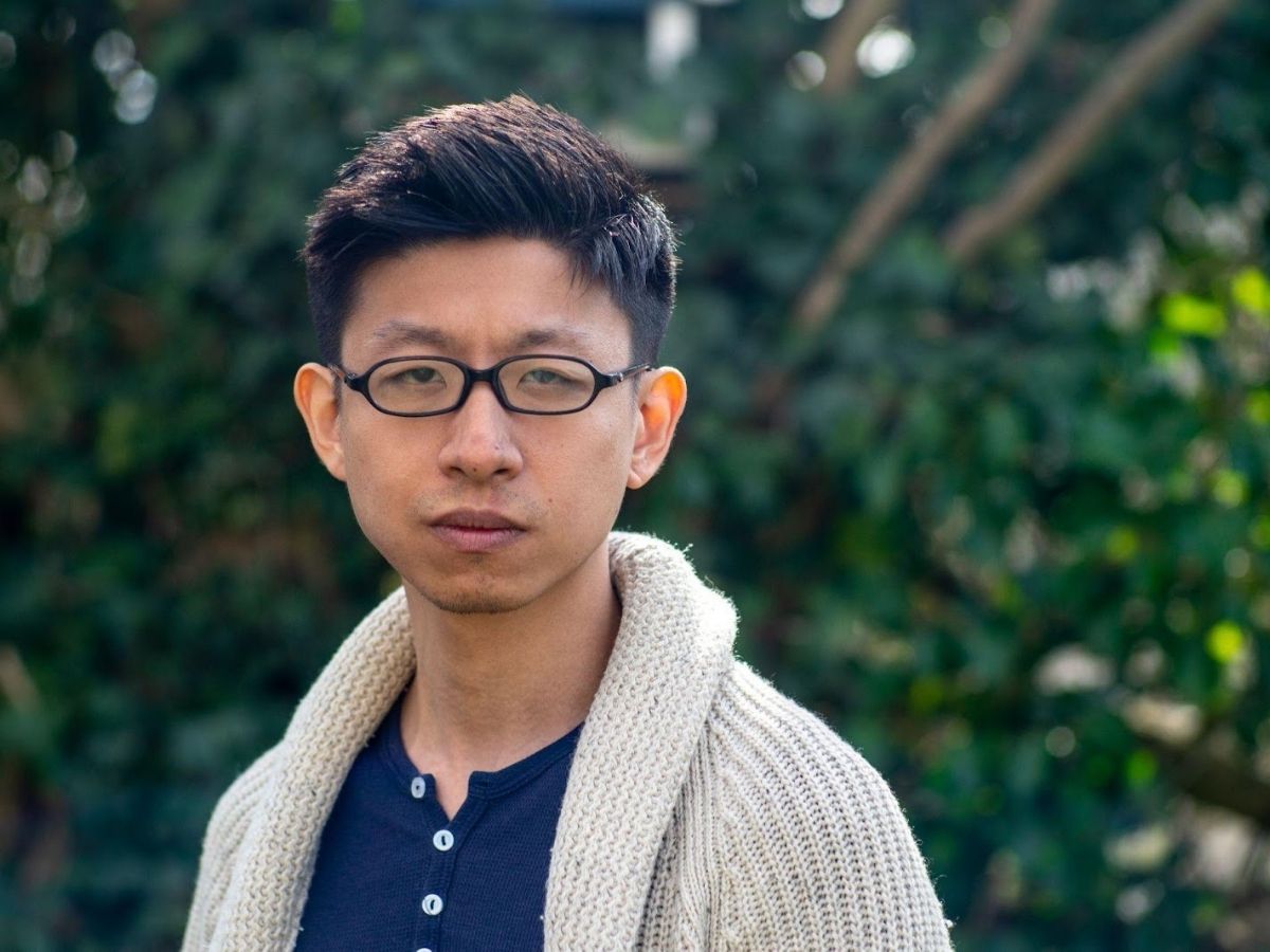 Author Ethan Lou wearing glasses, a beige cardigan and a blue undershirt standing in front of a wall of greenery.