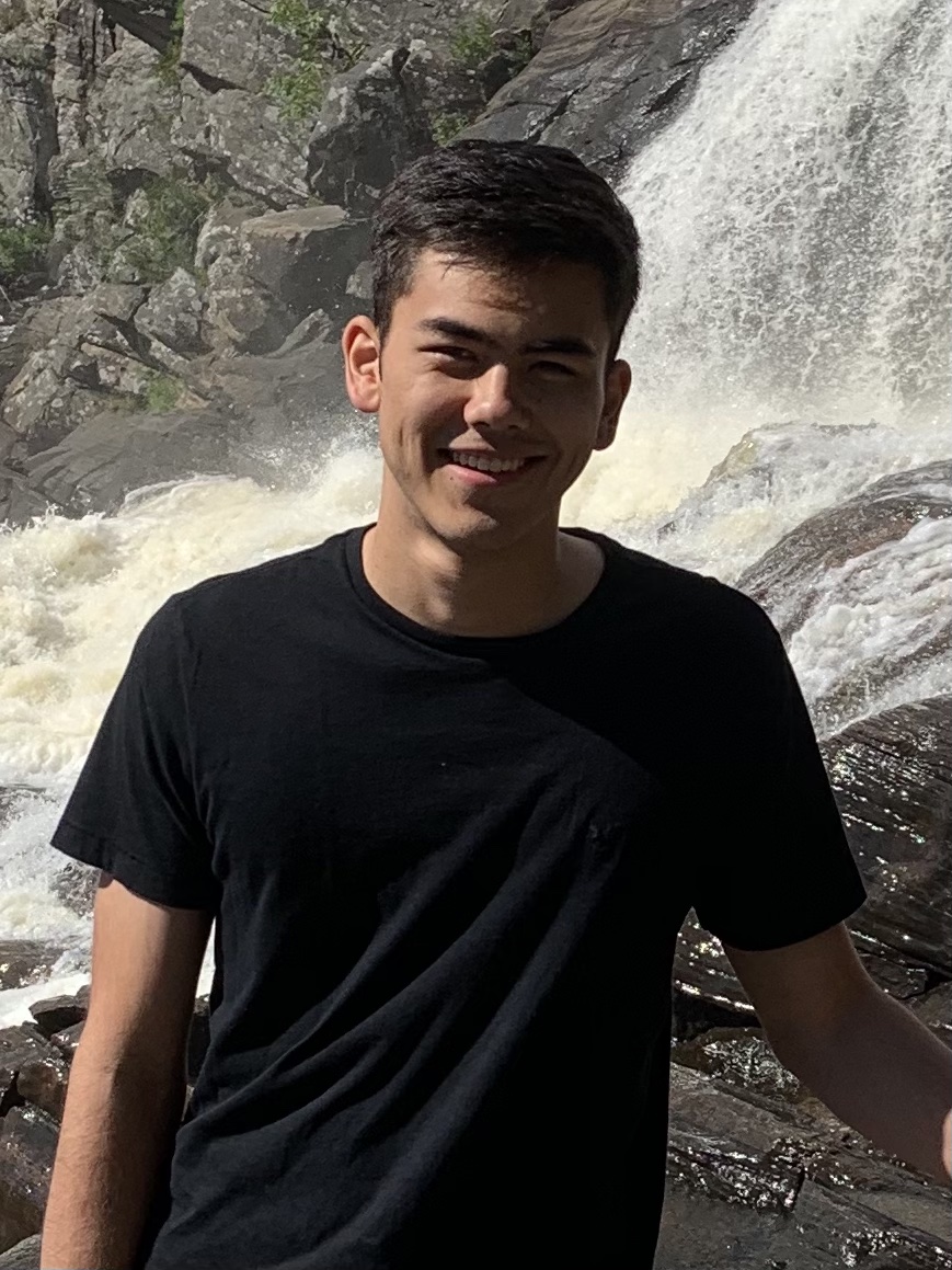 Evan, a young man with short black hair, wearing a black short sleeve shirt. He is standing in front of a waterfall, smiling.