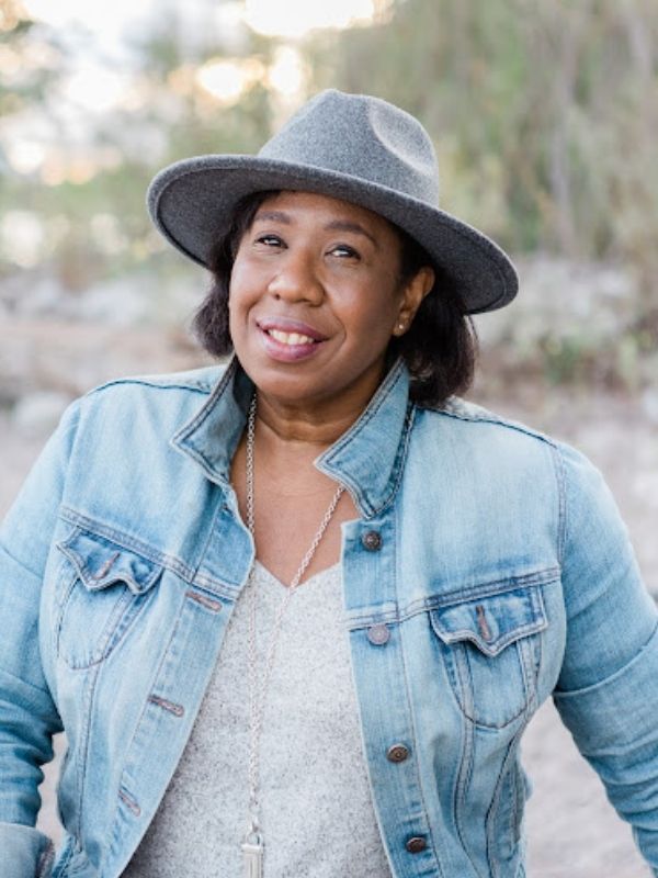 Margot Daley wearing a denim jacket over a grey shirt and a stylish grey hat standing in front of a blurred nature background